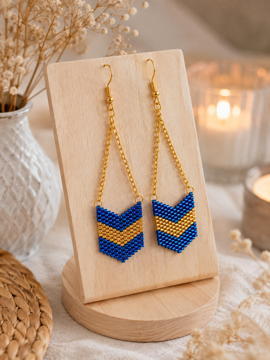 Paire de boucles d’oreilles Miyuki bleu irisé et doré présentée sur un support à bijoux en bois.