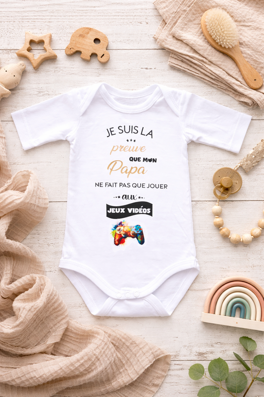 Body bébé blanc avec inscription humoristique sur papa gamer posé à plat avec accessoires bébé en décoration