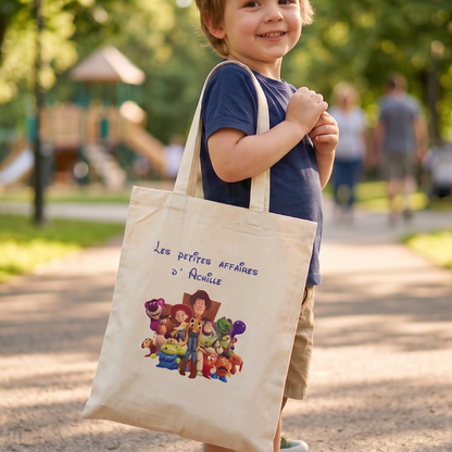 Tote bag enfant personnalisé avec prénom, sac en coton écru porté à l’épaule par un jeune enfant et illustré d’un univers de jouets colorés.