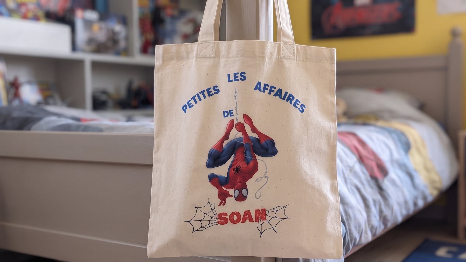 Tote bag enfant personnalisé avec prénom et illustration Spiderman, suspendu à un lit